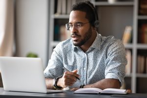 Focused young african businessman wear headphones study online watching webinar podcast on laptop listening learning education course conference calling make notes sit at work desk