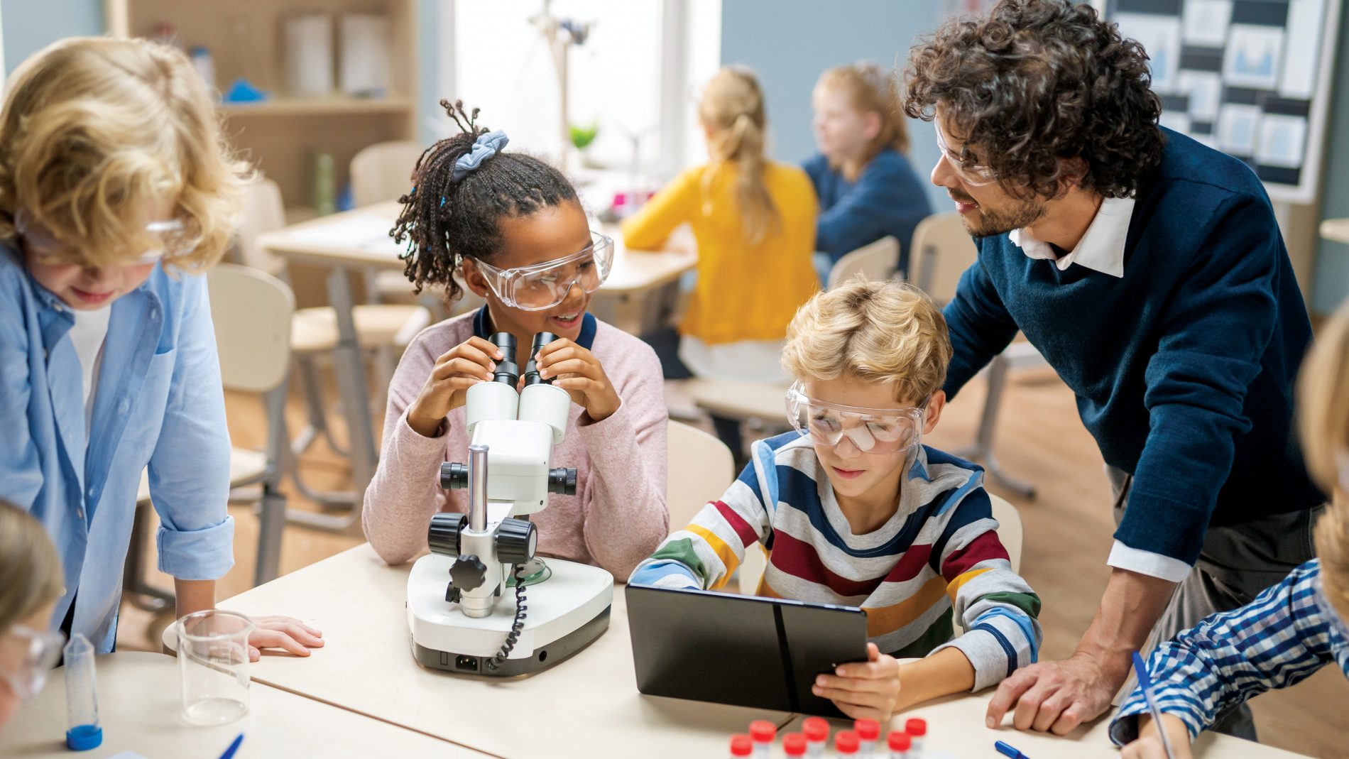 Elementary School Science Classroom: Cute Little Girl Looks Under Microscope, Boy Uses Digital Tablet Computer to Check Information on the Internet. Teacher Observes from Behind.