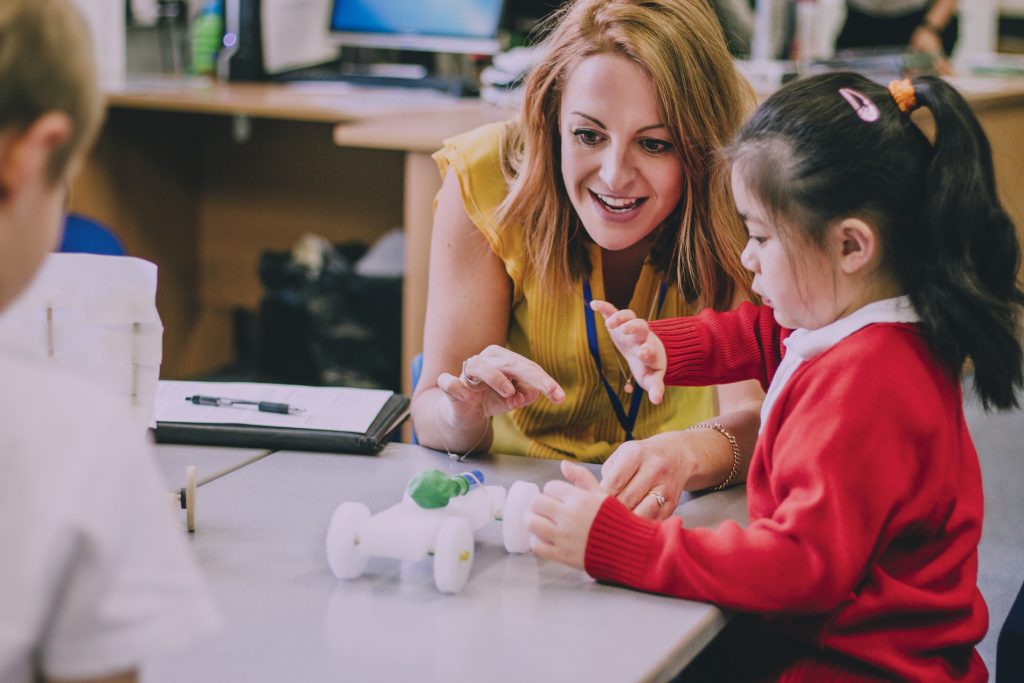 Teacher is sitting at the table in her classroom with her primary school students. They have built a car from recycled objects and crafts equipment and are testing that it works.