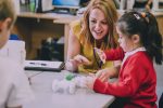 Teacher is sitting at the table in her classroom with her primary school students. They have built a car from recycled objects and crafts equipment and are testing that it works.